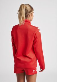 Rode sportieve jas met hoge kraag, voorzien van witte chevronaccenten op de schouders. Gecombineerd met bijpassende rode shorts, met een gladde textuur.
