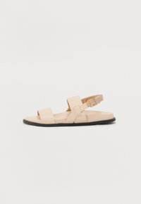 Sandale en cuir beige avec deux larges brides sur le pied et une bride de cheville ajustable, dotée d'une semelle en caoutchouc noir et d'un design minimaliste.
