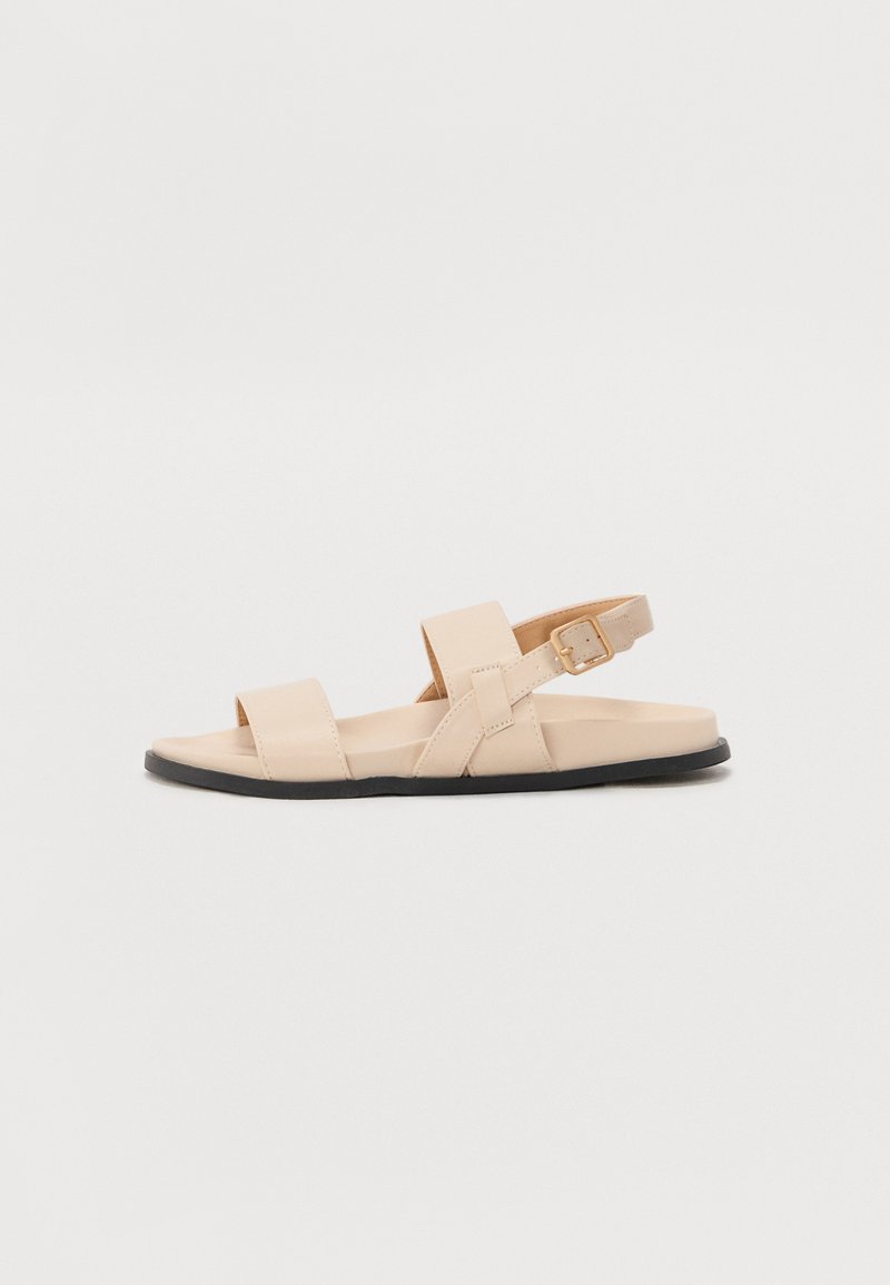 Sandale en cuir beige avec deux larges brides sur le pied et une bride de cheville ajustable, dotée d'une semelle en caoutchouc noir et d'un design minimaliste.