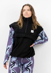 Schwarzer Fleece-Pullover mit hohem Kragen, Reißverschluss und Frontbranding. Getragen über einem langärmligen Oberteil im lila und blauen Camouflage-Muster und passenden Leggings.