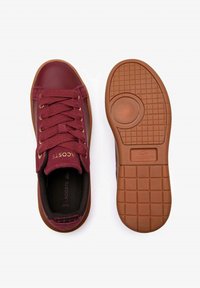 Sneaker bordeaux con tomaia in tessuto texture, occhielli dorati e suola in gomma marrone liscia con motivo a griglia. Logo Lacoste visibile.