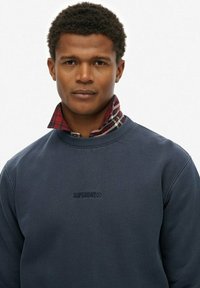 Marineblauwe sweater met reliëflogo, gelaagd over een kraag in rood en groen plaid. Soepele textuur, ronde halslijn en casual pasvorm.