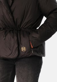 Svart dunjakke med innsvingt midje, med et gullemblem og sidelommer. Glatt, blank tekstur med en tettsittende silhuett.