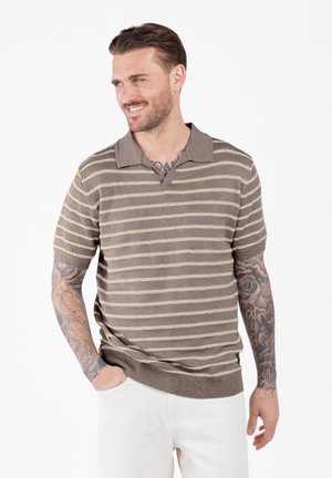 Hombre con tatuajes que lleva una camiseta polo de manga corta a rayas marrón y beige y pantalones blancos, sonriendo y mirando hacia un lado.