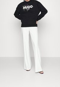 Zwarte sweatshirt met het logo "HUGO 1993" in kleurrijke print, gecombineerd met witte uitlopende broek en zwarte sandalen, staand tegen een effen achtergrond.