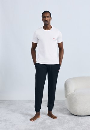 Bijela majica kratkih rukava s crvenim logotipom "HUGO", uparena s crnim teksturiranim trenirkom s crvenim logotipom, stilizirana s bosim nogama.