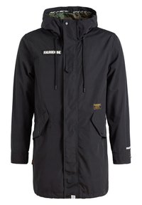 Parka noire longue à capuche avec poches à rabat sur le devant, cordons de serrage, et plusieurs petites écussons de marque sur la poitrine et la taille.