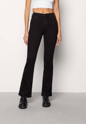 Kvinde iført sorte højttaljede flare jeans, hvid cropped top og sorte chunky sko, stående mod en ensartet lys baggrund.