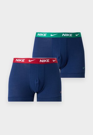 Homme torse nu portant un boxer bleu marine avec une ceinture rouge Nike, debout pieds nus sur un fond blanc uni.