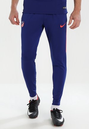 Beine in blauen Atletico Madrid Trainingshosen mit orangefarbenem Nike-Logo und schwarzen Fußballschuhen mit weißen und roten Details.