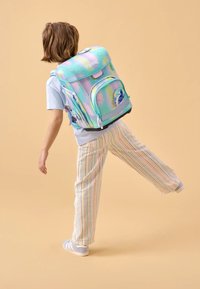 Sac à dos aux couleurs pastel arc-en-ciel, avec une finition brillante, un motif de bulles, une poche avant et des bretelles réglables. Enfant portant un pantalon rayé.