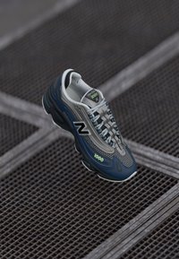 Plavi i sivi New Balance 1000 tenisice s crnim "N" logotipom suspendiranim iznad metalne rešetkaste podloge.