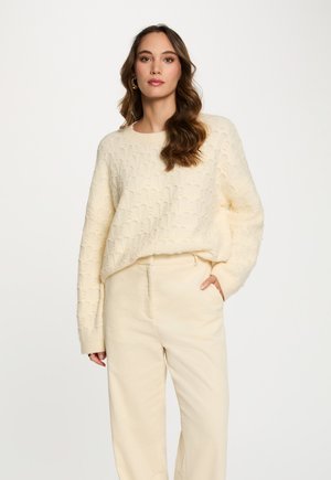 Donna con lunghi capelli mossi indossa un maglione testurizzato color crema e pantaloni a gamba larga abbinati, in piedi con una mano nella tasca su uno sfondo bianco.