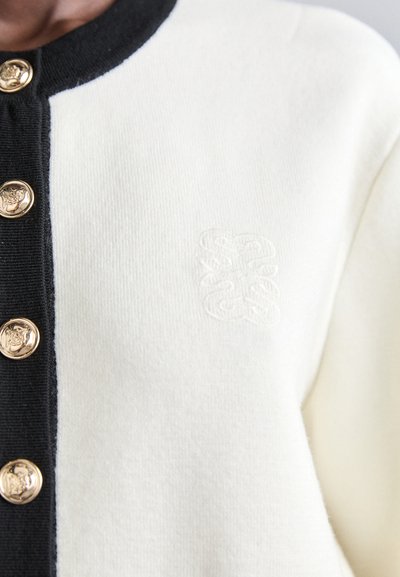 Gilet en tricot avec des blocs de couleur noire et crème, doté de boutons dorés et d'un logo décoratif en relief sur la zone gauche de la poitrine.