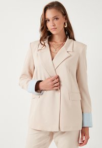 Bežový blazer s jednobunýkovým designem, klopovými límci a světle modrými žebrovanými manžetami. Dvě přední kapsy a hladká textilie.