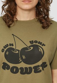 Olijf groene katoenen T-shirt met een zwarte afbeelding van twee kersen en de tekst "own your POWER" in vette letters. Ronde halslijn, relaxed fit.