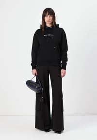 Felpa nera con cappuccio e tasca frontale, con testo bianco, abbinata a pantaloni neri a gamba larga con tasche laterali. Tiene una piccola borsa scura.