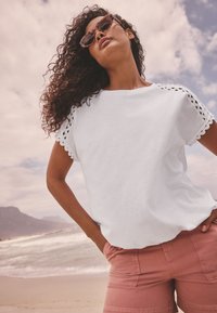 Kvinna med lockigt hår som bär solglasögon, en vit kortärmad topp med spetsdetaljer och rosa shorts, stående på en strand under en molnig himmel.