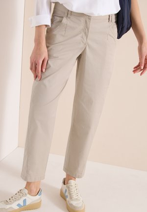 Person iført beige bukser med lige ben, hvid skjorte med optrukne ærmer og hvide sneakers med blå detaljer, stående på en lys gulv.