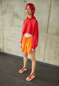 Röd kort hoodie med orange detaljer, kombinerad med orange och rosa shorts med dragsko. Bär röda och vita sneakers med guld detaljer.