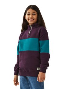 Leende flicka med långt hår som har på sig en lila och turkos tröja med halvmeterdragkedja och blå jeans, stående mot en vit bakgrund.