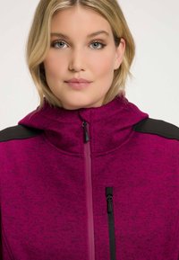 Fuchsia rits-hoodie met een textuurpatroon, capuchon en zwarte schouderaccenten. Voorzien van een voorzak met een zwarte rits.