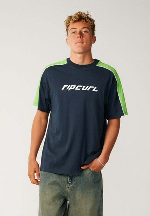 Joven con cabello rizado que lleva una camiseta Rip Curl azul marino y verde y vaqueros azul claro, de pie con una mano en el bolsillo.
