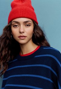 Maglione a righe navy con linee blu, sovrapposto a una camicia rossa. Il modello indossa un berretto di lana lavorato a maglia di un rosso vivace. Texture sottile e design casual.