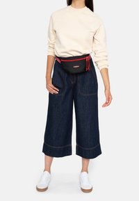 Black fanny pack with red accents worn over a cream sweatshirt and loose, cropped dark denim pants, paired with white sneakers.
