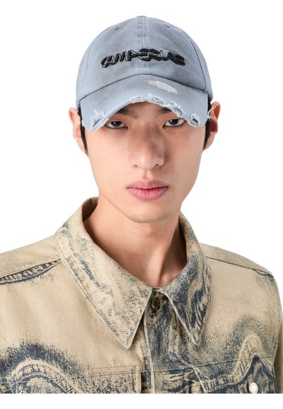 Light blue cap with distressed edges, featuring black embroidered text. Worn with a tan denim jacket showcasing a textured pattern.