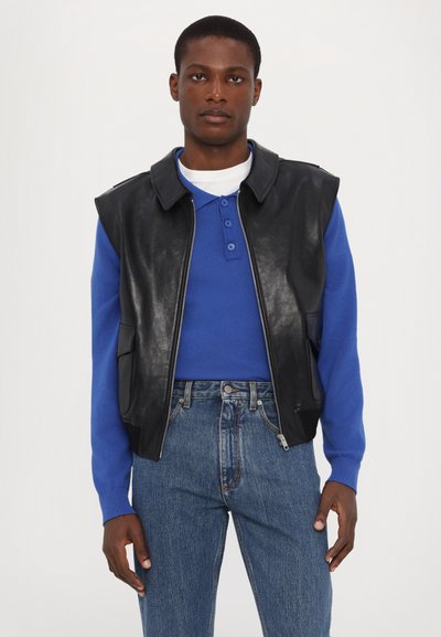 Black leather sleeveless jacket with a zip, worn over a blue long-sleeve shirt and light blue jeans. Features a classic collar and side pockets.