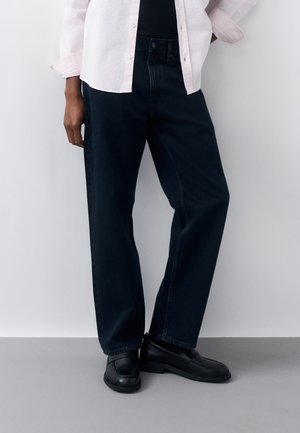 Person trägt lockere schwarze Jeans, schwarze Schuhe und ein hellfarbenes Hemd mit hochgekrempelten Ärmeln und steht vor einem schlichten Hintergrund.