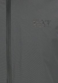 Svart zip-up jakke laget av teksturert stoff, har høy krage, vertikal glidelås og EA7-logo preget på brystet.
