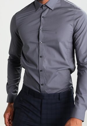 Mężczyzna w dopasowanej ciemnoszarej koszuli na guziki wpuszczonej do czarnych eleganckich spodni, stojący z częściowo widoczną jedną ręką.
