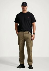 Mann iført svart caps og t-skjorte, khaki cargo-bukser, svart belte og klokke, som står med hendene i lommene foran en lys bakgrunn.