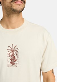 Camiseta de algodón color crema con un diseño gráfico marrón que presenta una serpiente y hojas de palma, el texto dice "BALANCE" y "OPPOSITES RVCA."