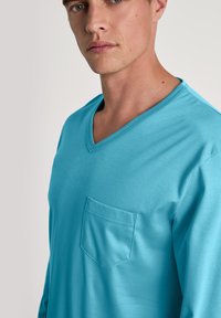 Turquoise V-hals shirt gemaakt van zachte stof, met een linkse borstzak en een lichtgewicht ontwerp, weergegeven op een model in profiel.