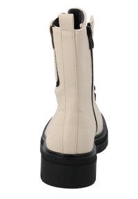 Bota de tobillo beige con suela negra, parte superior sintética texturizada, cremallera lateral y acentos en negro contrastantes para un diseño moderno.