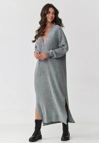 Długa, szara, prążkowana dzianinowa sukienka z głębokim dekoltem w kształcie V, długimi rękawami i bocznymi rozcięciami, w zestawieniu z czarnymi butami na sznurówki.