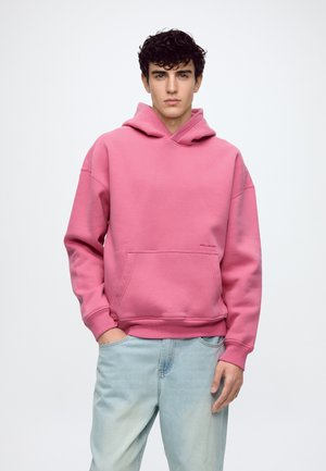 Jeune homme aux cheveux bouclés et foncés, portant un sweat à capuche rose et un jean bleu clair, debout avec une main dans la poche, sur un fond uni.