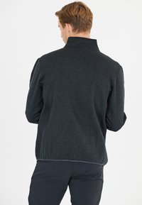 Giacca pullover nera, texturizzata, con collo alto e zip sul retro; presenta cuciture piatte e una vestibilità aderente, progettata per comfort e mobilità.
