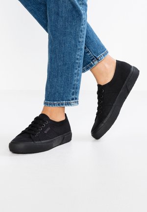 Person trägt schwarze Low-top-Sneaker und blaue Jeans und steht auf einem weißen Boden mit einem leicht angehobenen Fuß.