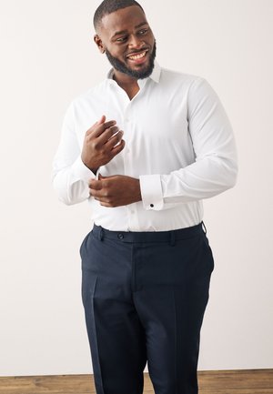 Cămașă albă cu nasturi, cu mâneci lungi și butoni negri, asortată cu pantaloni de costum de un albastru închis, având un material neted și un design ajustat.