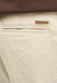 Biege kokvilnas bikses ar aizmugurējo kabatu, kas aprīkota ar horizontālu atvērumu, un nelielu brūnu ādas etiķeti ar iegravētu "JACK & JONES".