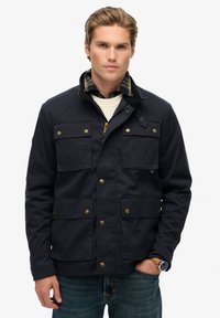 Marineblauwe jacket van stevig materiaal, met vier voorzakken, messing knopen en een kraag met daaronder een gestreept shirt.