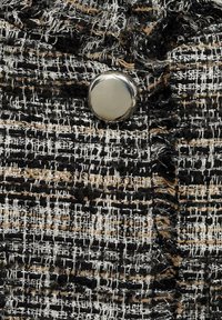 Czarnobiały, teksturowany materiał z beżowymi akcentami, z błyszczącym srebrnym guzikiem z przodu. Wzór tkany z postrzępionymi krawędziami.