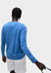 Pánský modrý pletený svetr s dlouhými rukávy a žebrovaným lemem, nošený přes bílé lněné šortky, který ukazuje hladkou texturu a uvolněný střih.