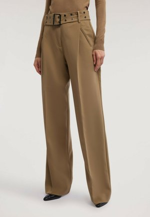 Mujer con pantalones anchos de talle alto en color camello, con cinturón a juego y bolsillos laterales con cremallera, combinados con zapatos negros puntiagudos.