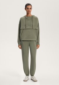 Olivově zelená mikina s klokaní kapsou a stahovacími šňůrkami, spárovaná s odpovídajícími zúženými tepláky. Tenisky v světle šedé a bílé.