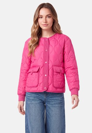 Kvinde med langt hår iført en knaldrød quiltet jakke med trykknapper og klaplommer, kombineret med blå jeans, stående mod en hvid baggrund.